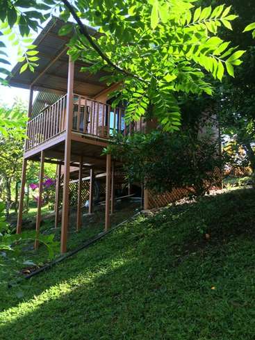 The image depicts a two-story wooden house with a balcony, situated on a hillside surrounded by lush greenery and trees, bathed in sunlight.