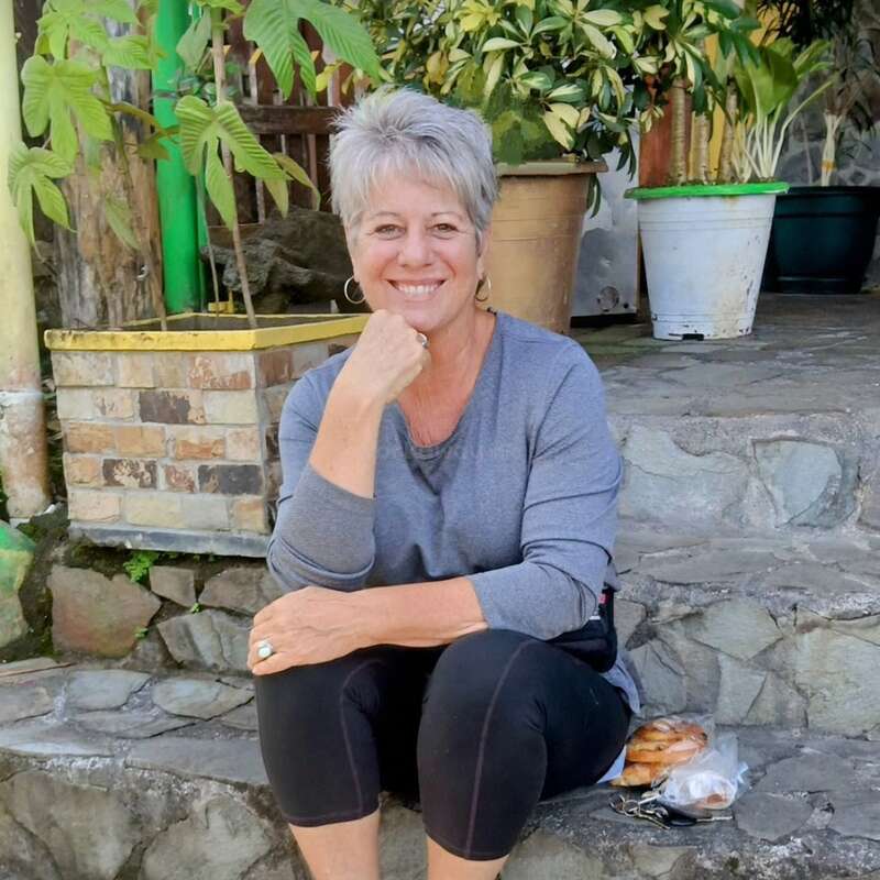 The image depicts a woman sitting on stone steps, with short grey hair, wearing a grey shirt and black pants, surrounded by potted plants and a bag of food nearby.