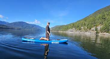 A man kneels on a blue paddleboard in calm lake water, surrounded by green forested hills and mountains under a bright, clear blue sky.
