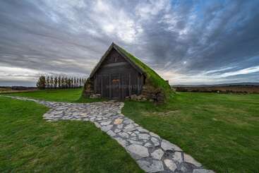 Una pequeña y rústica casa de césped con tejado verde se asienta sobre un terreno abierto cubierto de hierba, un camino de piedra conduce a su puerta bajo un espectacular cielo nublado.