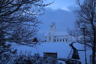 Una iglesia blanca se alza apaciblemente rodeada de nieve, bajo un cielo invernal teñido de azul. Árboles cubiertos de nieve enmarcan la serena escena, con un camino despejado que conduce hacia delante.