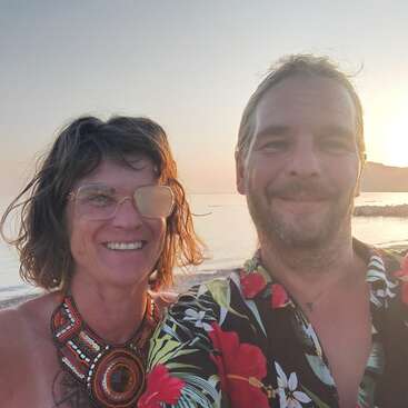 A smiling couple poses by the beach at sunset. The woman wears glasses with one lens covered and a bold necklace; the man wears a floral shirt.