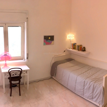 This cozy, minimalist bedroom features a single bed, a floating shelf with books, a small desk with a pink lamp, a chair, and a bright window.