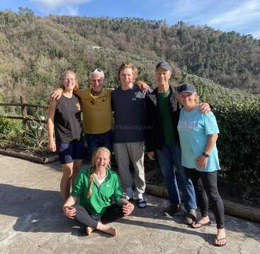 A imagem mostra um grupo de seis pessoas posando para uma foto em um pátio de pedra, em um cenário pitoresco de uma colina arborizada sob um céu azul com nuvens brancas.