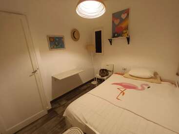 A cozy bedroom featuring a flamingo bedspread, wall art, a hat, a lamp, a chair with a cat, white walls, and a shelf with decorations.
