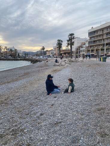 Two people sit on a pebbled beach by the sea, surrounded by palm trees, buildings, and a cloudy sky at sunset, creating a peaceful atmosphere.