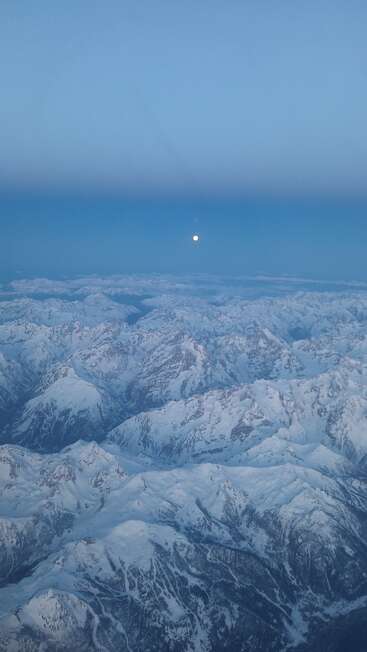 This image captures a breathtaking aerial view of snow-covered mountains beneath a clear blue sky, with the full moon shining brightly above the stunning landscape.