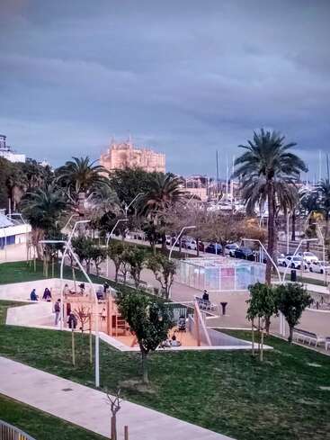 A park features a playground with children and adults, surrounded by palm trees. In the background, there are docked boats and a historic cathedral.