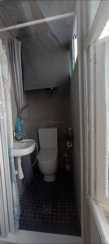 This image shows a very narrow bathroom with a toilet, small sink, handheld shower, and tiled floor. There’s a curtained window and minimal space available.