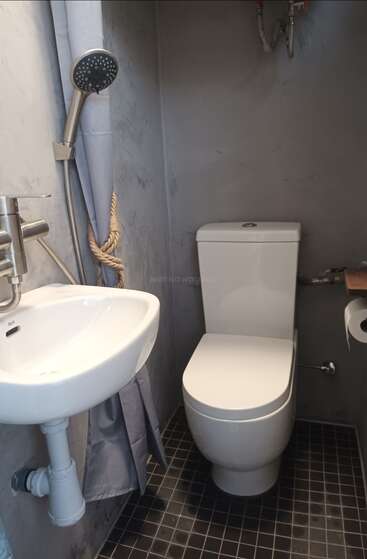 This small modern bathroom features a white toilet, compact sink with faucet, handheld showerhead, gray tiles, and minimal decor. A rope curtain tie adds rustic charm.