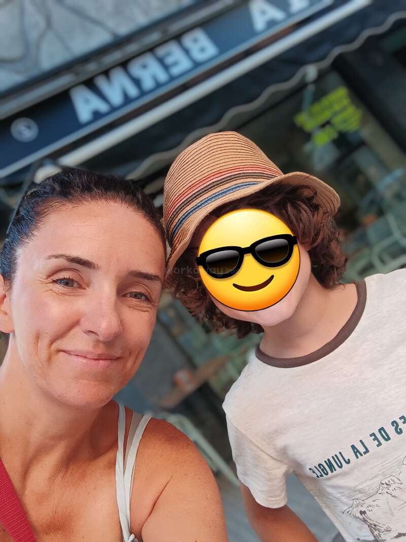 Das Bild zeigt eine Frau und ein Kind, die für ein Selfie posieren, wobei das Gesicht des Kindes von einem Emoji verdeckt wird. Die Frau trägt ein weißes Tank-Top.