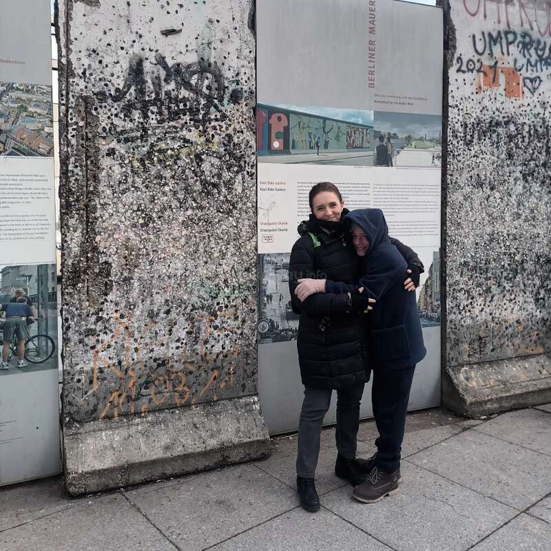 Two people warmly hugging stand beside graffiti-covered Berlin Wall remnants. Informational panels describe the historical site. It's a cold day, and both wear jackets, smiling gently.