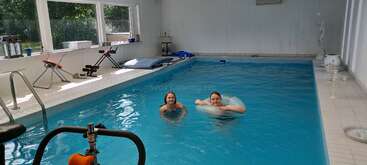 Deux personnes nagent dans une piscine intérieure, l'une d'elles utilisant un anneau de flottaison. Des équipements d'exercice et de grandes fenêtres sont visibles, permettant à la lumière naturelle de pénétrer dans la pièce.