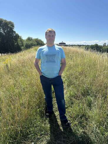 Un homme, vêtu d'un t-shirt bleu "GENIUS" et d'un jean, se tient debout dans un champ herbeux éclairé par le soleil, avec des arbres et un bâtiment à l'arrière-plan.