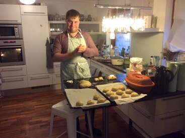 Dans une cuisine moderne, un homme en tablier façonne des boules de pâte, tandis que des plateaux de petits pains non cuits sont posés sur le comptoir à côté de lui.