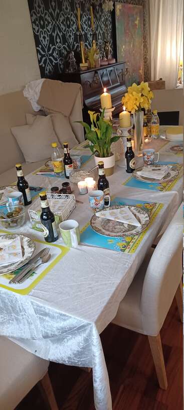 Une table à manger festive avec des décorations florales, des bougies, des assiettes, des ustensiles, des bières, des tasses et des jonquilles. Des sets de table joyeux et des sièges confortables créent une atmosphère accueillante.