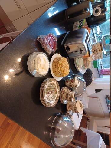 Une table de petit-déjeuner avec des gaufres, de la crème fouettée, de la confiture, un grille-pain, une bouilloire électrique, des assiettes empilées, des tasses et un plat couvert, le tout disposé sur un plan de travail sombre.