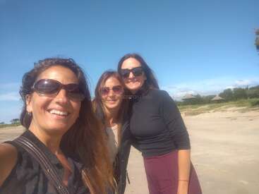 Drei Frauen stehen zusammen an einem sonnigen Strand, alle tragen Sonnenbrillen und lächeln, mit blauem Himmel, Sand und Grün im friedlichen Hintergrund.
