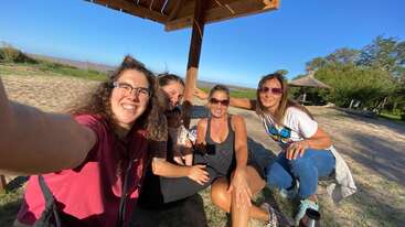 Quatre femmes sont assises et sourient sous une structure en bois par une journée ensoleillée. Elles se trouvent en plein air, dans un parc, et profitent ensemble de leur compagnie, de leurs rires et de la nature.