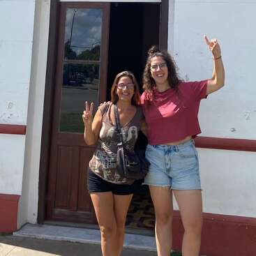 Deux femmes se tiennent debout devant un immeuble, toutes deux souriantes, faisant le signe de la paix. L'une porte un haut court rouge et un short en jean, l'autre un débardeur à motif et des lunettes.