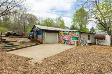 Ein bescheidenes Haus mit buntem Graffiti auf dem Garagentor, umgeben von Bäumen, Holzspänen und Gegenständen im Freien, mit einer Betonauffahrt und einem rustikalen Hof.