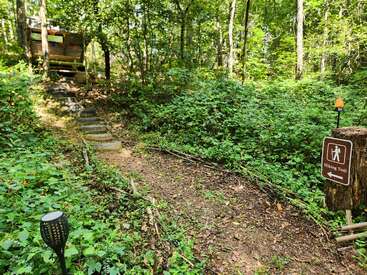 Ein unbefestigter Wanderweg schlängelt sich durch einen üppigen grünen Wald. Ein Schild weist den Weg. Holzstufen führen hinauf zu einer Hütte, die teilweise von oben zu sehen ist.