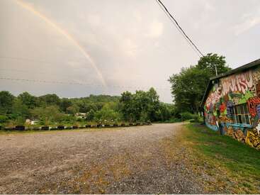 Ein schwacher Regenbogen wölbt sich über eine ländliche Szene mit üppig grünen Bäumen und Hügeln, einer Schotterstraße und einem Gebäude mit bunten Graffiti auf der rechten Seite.