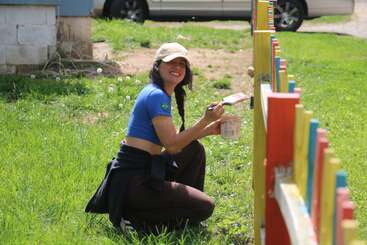Eine junge Frau kniet lächelnd im Gras und streicht einen bunten Holzzaun. Sie trägt ein blaues Hemd, eine braune Hose und eine helle Mütze. Ein sonniger Tag.