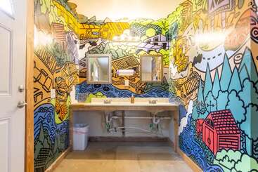 Dieses Badezimmer ist mit farbenfrohen Wandbildern im Cartoon-Stil ausgestattet, die Häuser, Bäume und Flüsse darstellen. Es hat zwei Waschbecken, darunter freiliegende Rohrleitungen, zwei Spiegel und eine helle Beleuchtung.