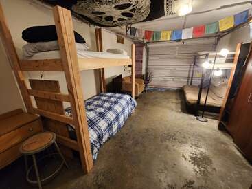 Dieses Bild zeigt ein behelfsmäßiges Schlafzimmer in einer Garage mit Etagenbetten, einem Futon, bunten Fahnen, Betonböden, bescheidenen Möbeln und einem Wandteppich an der Decke.