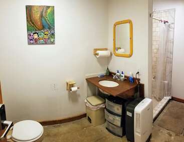 Dieses Badezimmer verfügt über eine Toilette, ein Eckwaschbecken mit gelbem Spiegel, eine Dusche mit Vorhang, zwei Abfalleimer, Stauschubladen, Wandkunst und einen weißen Luftentfeuchter.