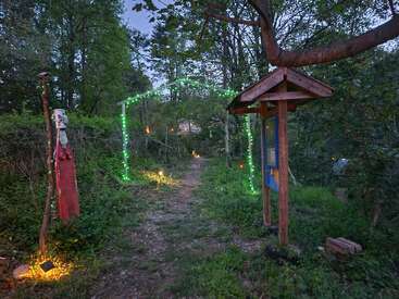 Ein Waldweg ist mit grünen Lichterketten, leuchtenden Laternen, einem rustikalen Torbogen, Holzskulpturen und einem kleinen überdachten Schild geschmückt und schafft eine magische Atmosphäre.