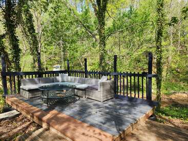 Eine gemütliche Außenterrasse mit einem grauen Sektionssofa und einem gläsernen Couchtisch liegt inmitten hoher Bäume, umgeben von üppigem Grün und friedlicher Natur.