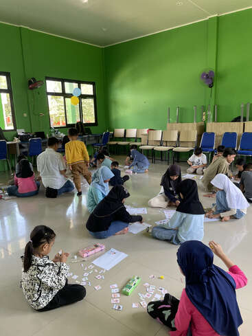 A group of children sit on the floor in a green-walled classroom, playing educational games with cards and papers. Chairs are stacked against the wall.