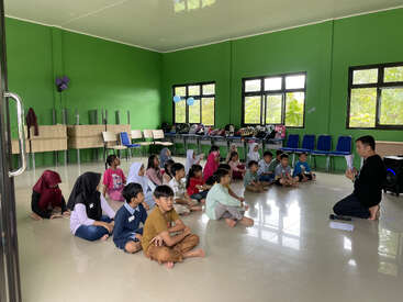 A group of children sit on the floor in a green classroom, attentively listening to an adult teacher. Chairs and backpacks line the back of the room.