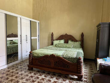 This cozy bedroom features an ornate wooden bed with green bedding, a mirrored white wardrobe, tiled floor, dark cabinet, and soft yellow walls, exuding warmth.