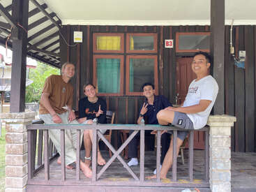 Four friends sit and smile on a wooden porch outside a house labeled B3, enjoying a relaxed moment together. Casual, friendly atmosphere with peace signs.