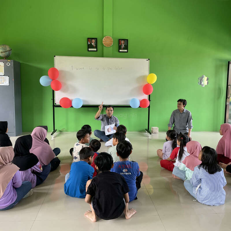 A teacher sits on the floor instructing a group of children, with another adult nearby. The classroom has a green wall, whiteboard, and colorful balloons.