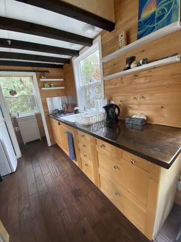 Cette cuisine confortable comprend des armoires en bois, des plans de travail foncés, des étagères ouvertes décorées, de grandes fenêtres laissant entrer la lumière naturelle, un parquet foncé, des poutres apparentes et un agencement compact.