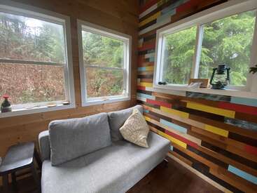 Un coin confortable avec un canapé gris clair orné d'un coussin à motifs, un mur en bois aux motifs géométriques colorés, de grandes fenêtres donnant sur une forêt verdoyante et une décoration rustique.