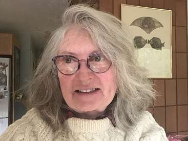 Une femme aux cheveux gris et portant des lunettes sourit légèrement. Elle porte un pull couleur crème. Derrière elle, on aperçoit des œuvres d'art accrochées au mur et un intérieur chaleureux et confortablement éclairé.