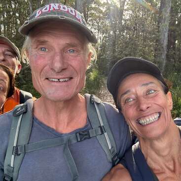 Four smiling adults, two in the foreground and two partially visible in the background, enjoy a sunny outdoor hike in a wooded forest, wearing casual hats and backpacks.