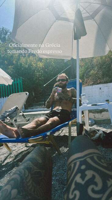A tattooed man relaxes on a lounge chair under an umbrella, sipping Fredo espresso in Greece; another tattooed person's legs are visible in the foreground.