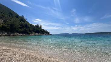 This serene coastal scene features clear turquoise water, a pebbled beach, lush green hills on the left, and a bright blue sky with scattered clouds.