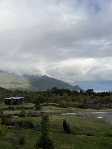 A small cabin stands in a lush, green landscape with a river, trees, and distant mountains. A faint rainbow appears under dramatic, cloudy skies. Peaceful scenery.