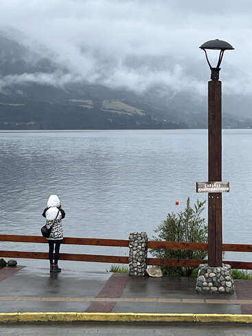 A person in a white jacket stands by a lakeside railing, facing misty mountains. A streetlamp and sign are nearby. The atmosphere is calm and overcast.