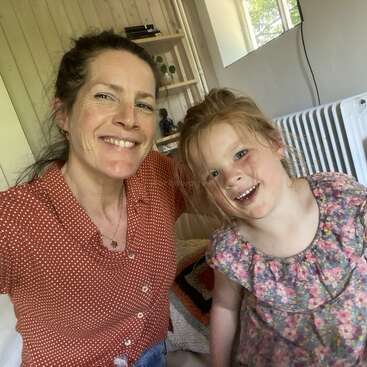Uma mulher e uma jovem sorridentes posam para uma selfie em um ambiente interno. O plano de fundo apresenta paredes de madeira clara, um radiador, uma janela e prateleiras com pequenos itens decorativos.