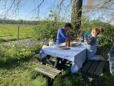 Um homem e uma criança desfrutam de uma refeição ao ar livre em uma mesa de piquenique coberta com um pano azul, cercada por vegetação, flores e a bela luz do sol da primavera.