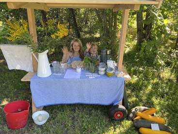 Duas crianças sorridentes acenam de trás de uma barraca caseira ao ar livre, decorada com flores, abobrinhas e vários potes, em um jardim verde e ensolarado entre árvores.