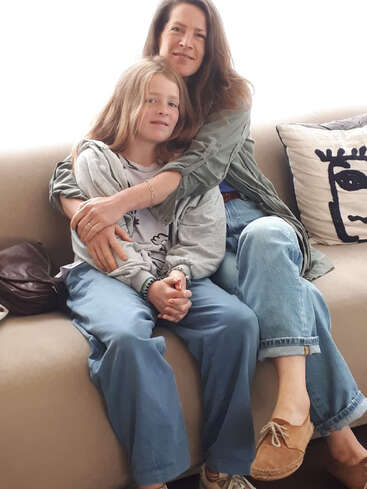 Uma mulher e uma criança sentam-se juntas em um sofá bege, abraçando-se carinhosamente. Ambos estão sorrindo gentilmente. Há um travesseiro decorativo e uma bolsa escura ao lado deles.